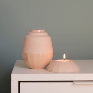Kleine urnenset in karamelkleur, bestaande uit een handgemaakte keramieken urn en een bijpassende waxinelichthouder met brandend kaarsje, geplaatst op een wit kastje tegen een zachtgroene muur.