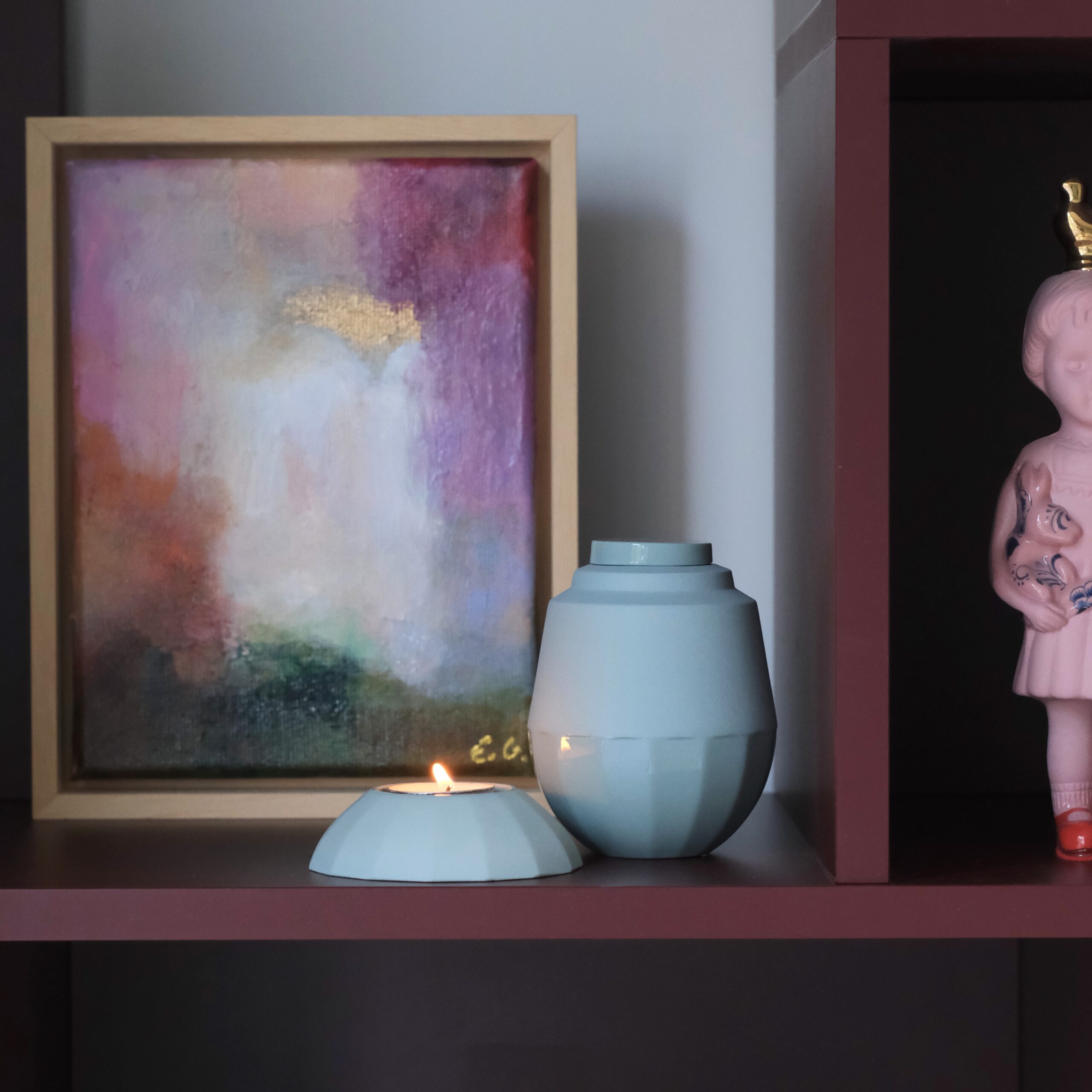 Kleine mintkleurige urnenset met waxinelichthouder en brandend kaarsje, stijlvol gepresenteerd op een bordeauxrode plank voor een abstract schilderij in warme tinten, in een houten lijst.