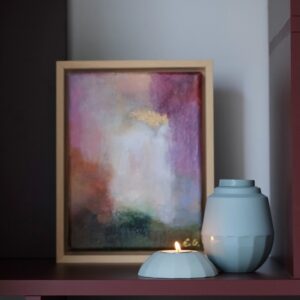 Kleine mintkleurige urnenset met waxinelichthouder en brandend kaarsje, stijlvol gepresenteerd op een bordeauxrode plank voor een abstract schilderij in warme tinten, in een houten lijst.